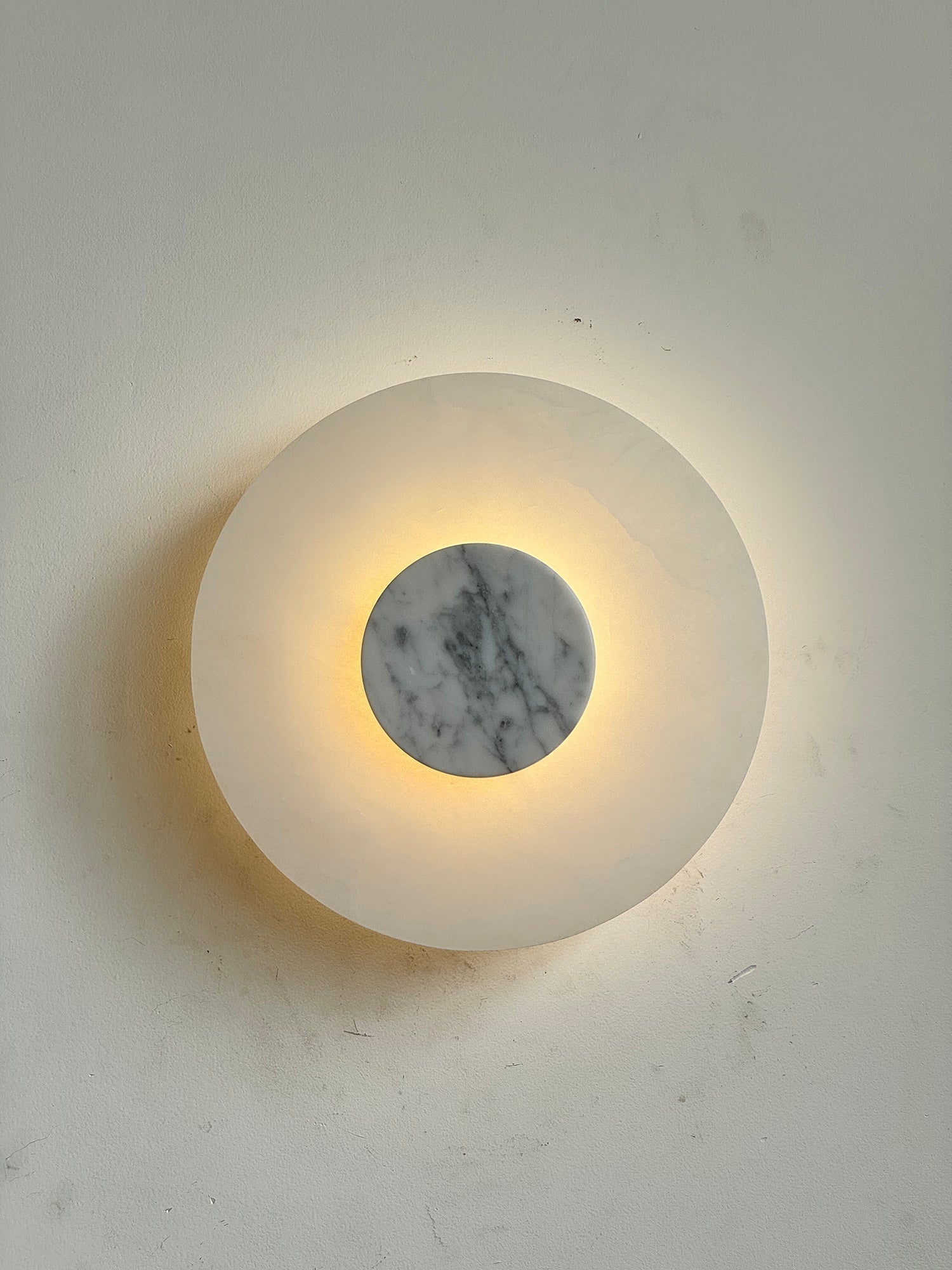 Clérachic rauduvyi wall lamp lit head-on, the alabaster plate glowing and casting a soft halo on the wall
