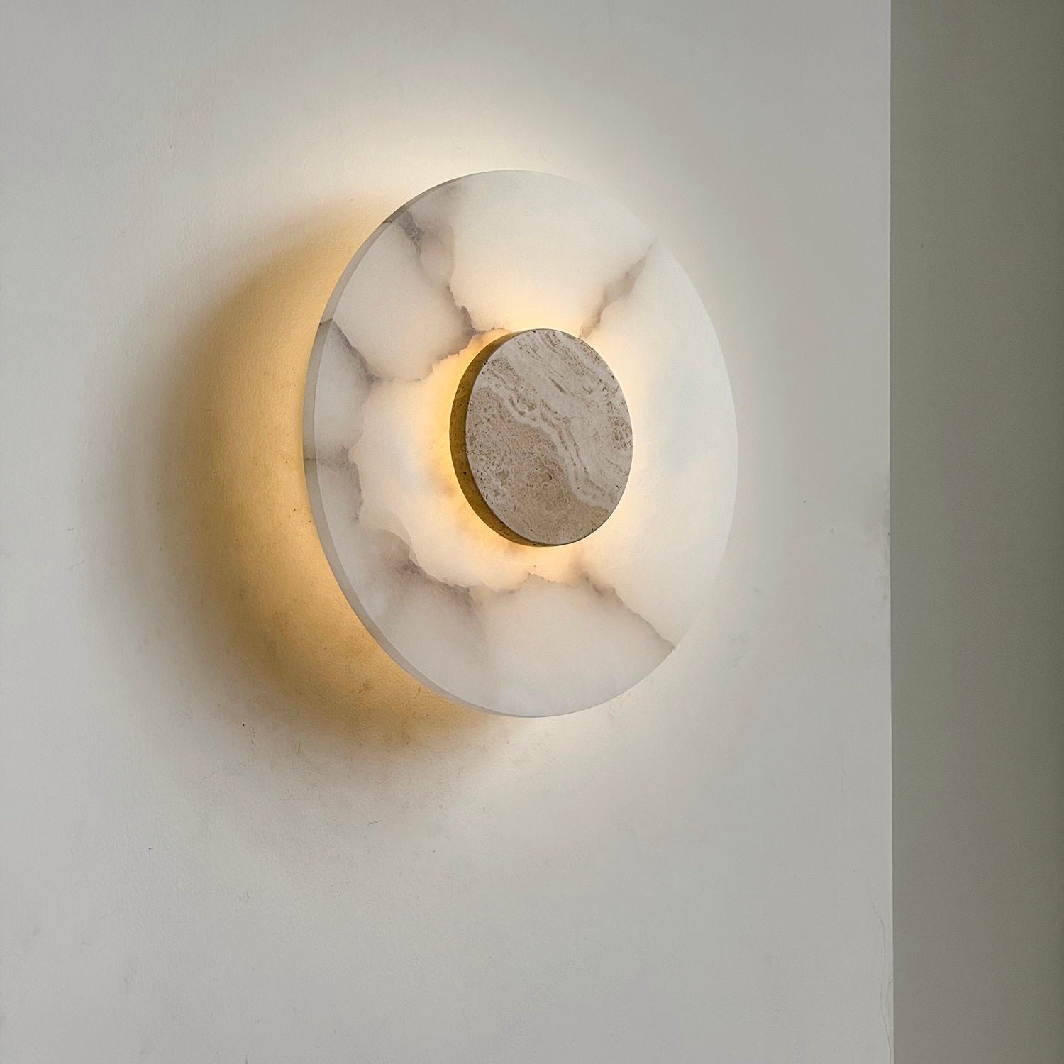 Clérachic rauduvyi wall lamp lit at an angle, the beige travertine disc framed by a halo of warm light