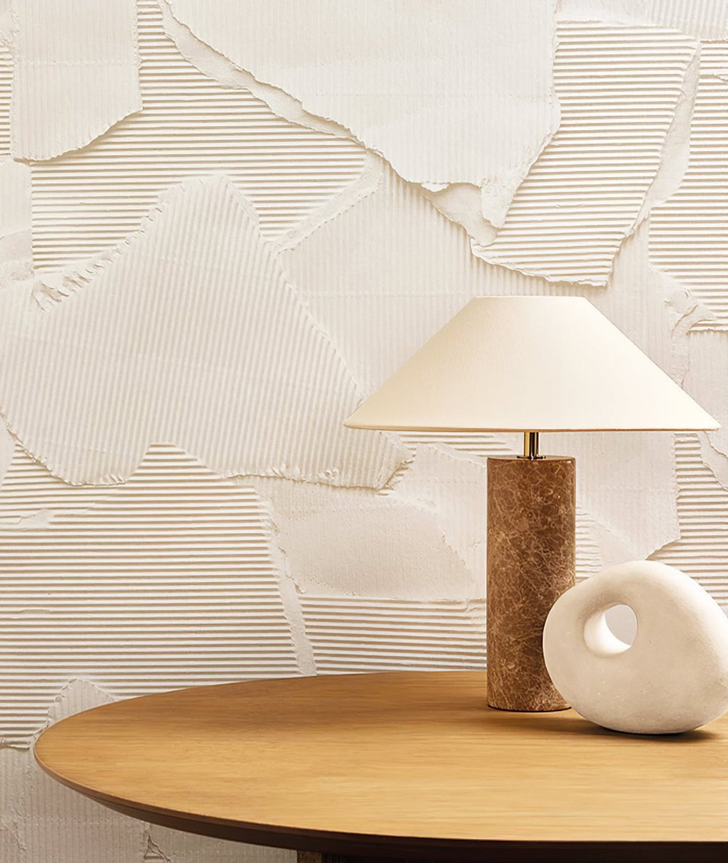 The Clérachic table lamp sits on a round wood side table against a textured plaster wall, beside a white pebble-shaped sculpture.