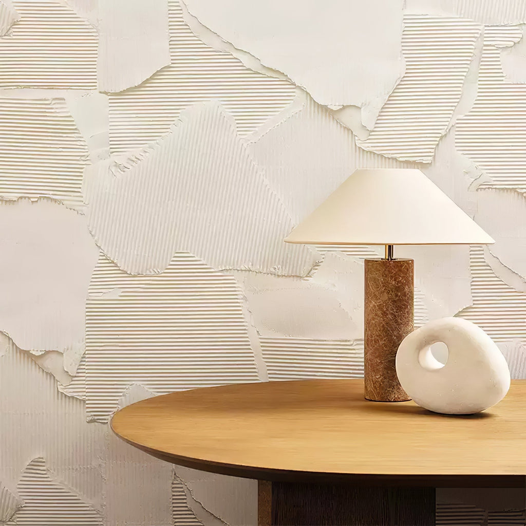 A wider view of the Clérachic table lamp on the round wood side table, the white pebble sculpture beside it against the plaster wall.