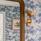 Gold-leaf Kristy sconce lit on blue botanical wallpaper next to a gilt-framed mirror.