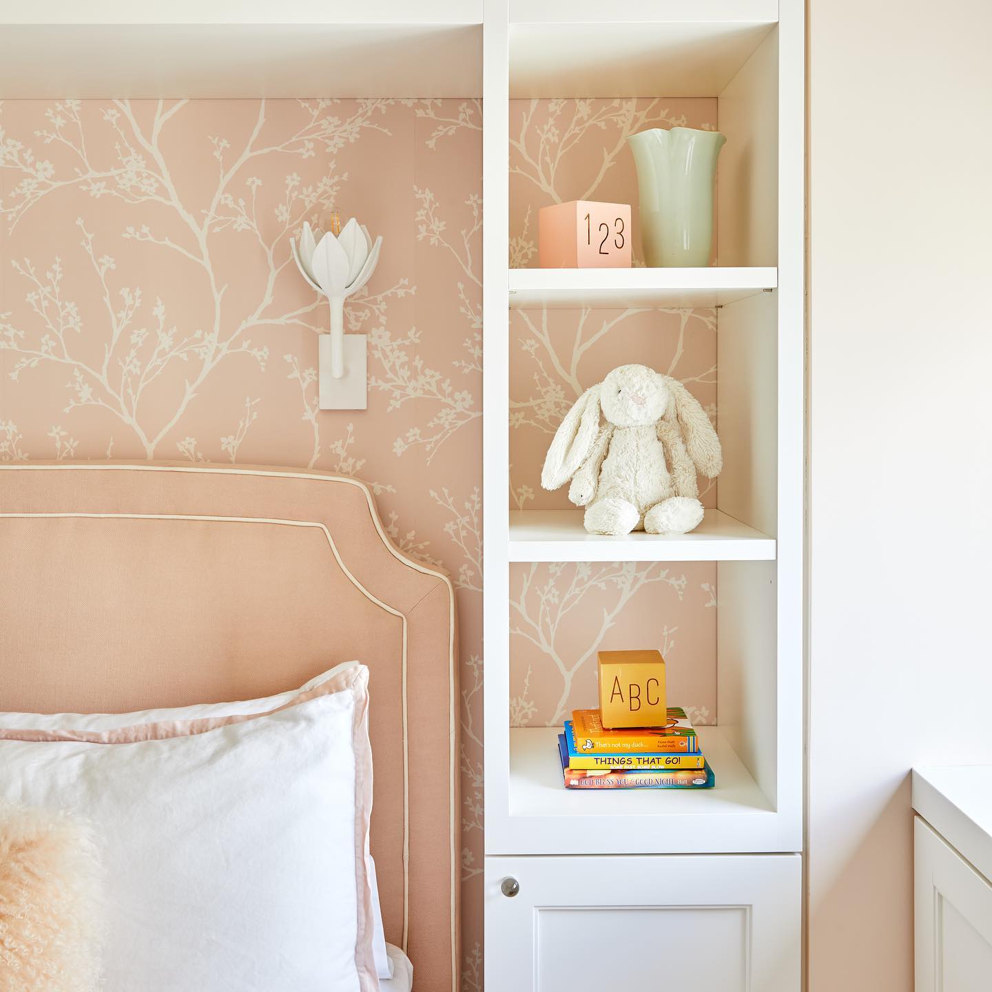 White plaster Kristy sconce above an upholstered headboard on blush blossom wallpaper in a child's room.