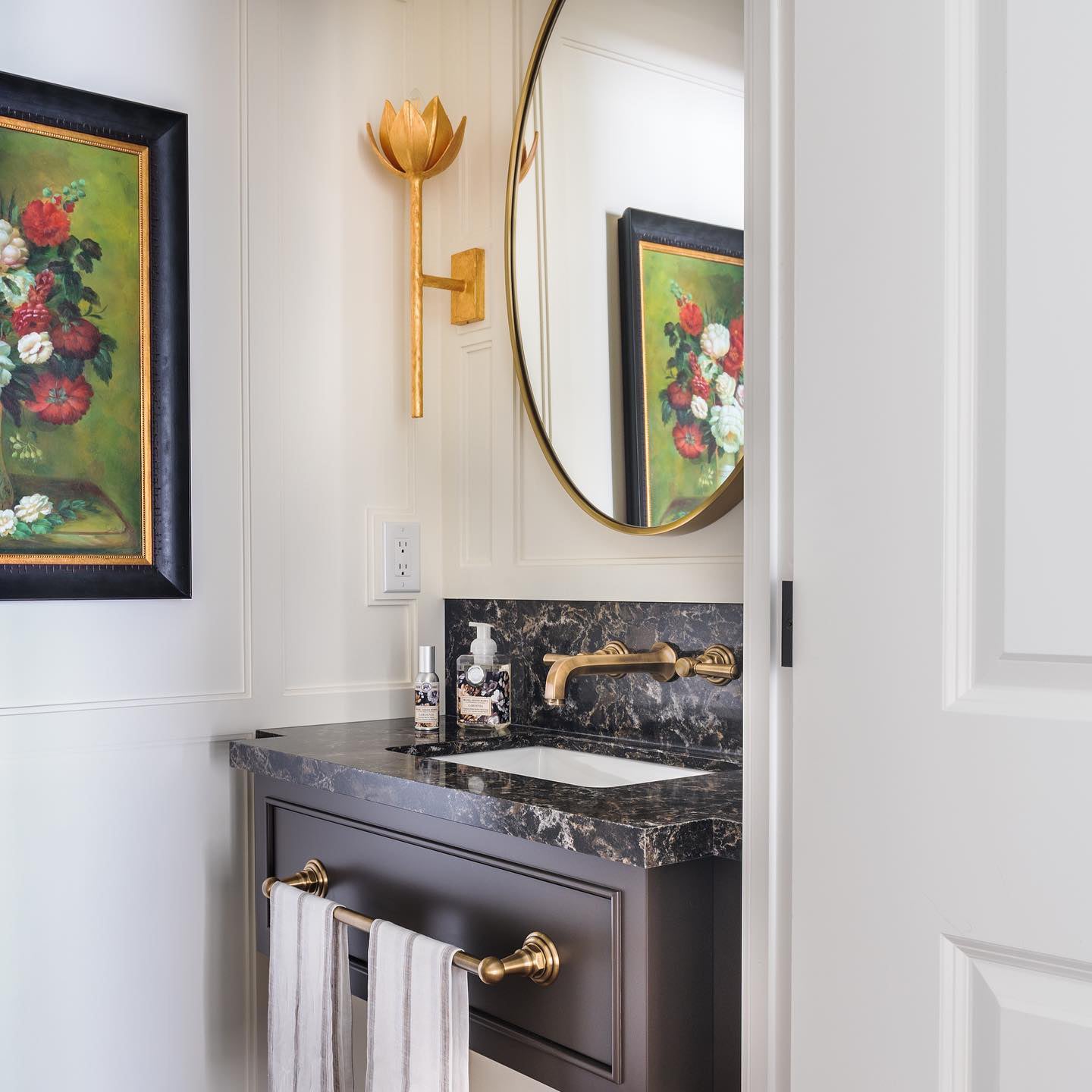 Gold-leaf Kristy sconce beside a round brass mirror in a panelled powder room with a marble vanity.