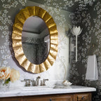 White Kristy sconce on olive blossom wallpaper across from a gold scalloped oval mirror and marble sink.