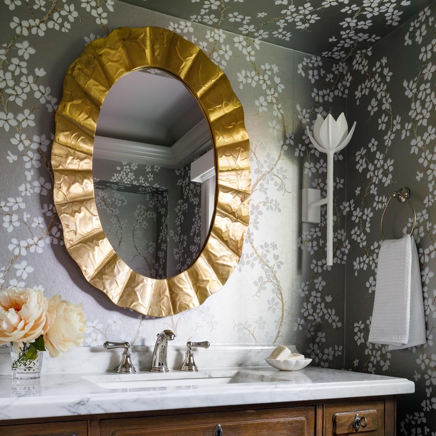 White Kristy sconce on olive blossom wallpaper across from a gold scalloped oval mirror and marble sink.