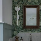 White plaster Kristy sconce on deep green floral wallpaper next to a dark wood framed bathroom mirror.