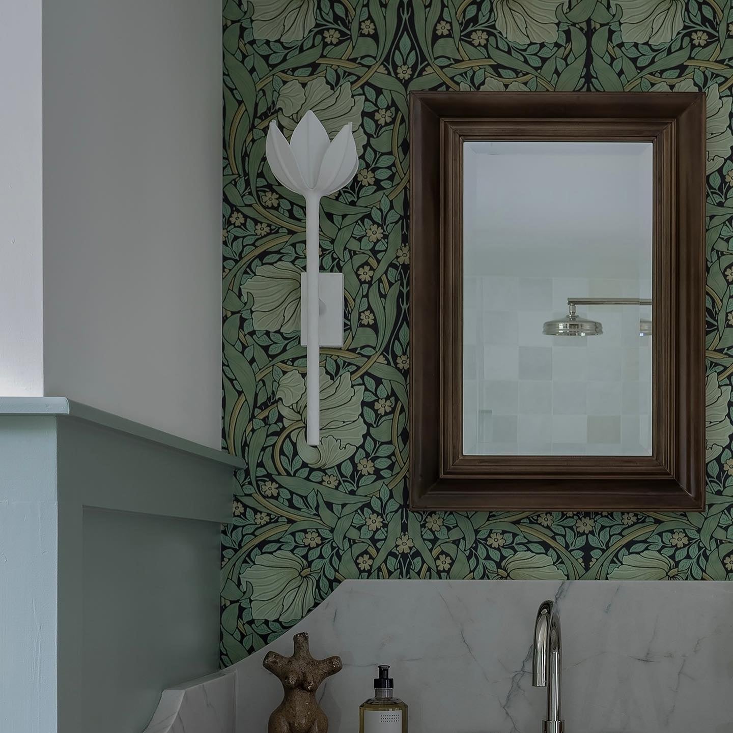 White plaster Kristy sconce on deep green floral wallpaper next to a dark wood framed bathroom mirror.