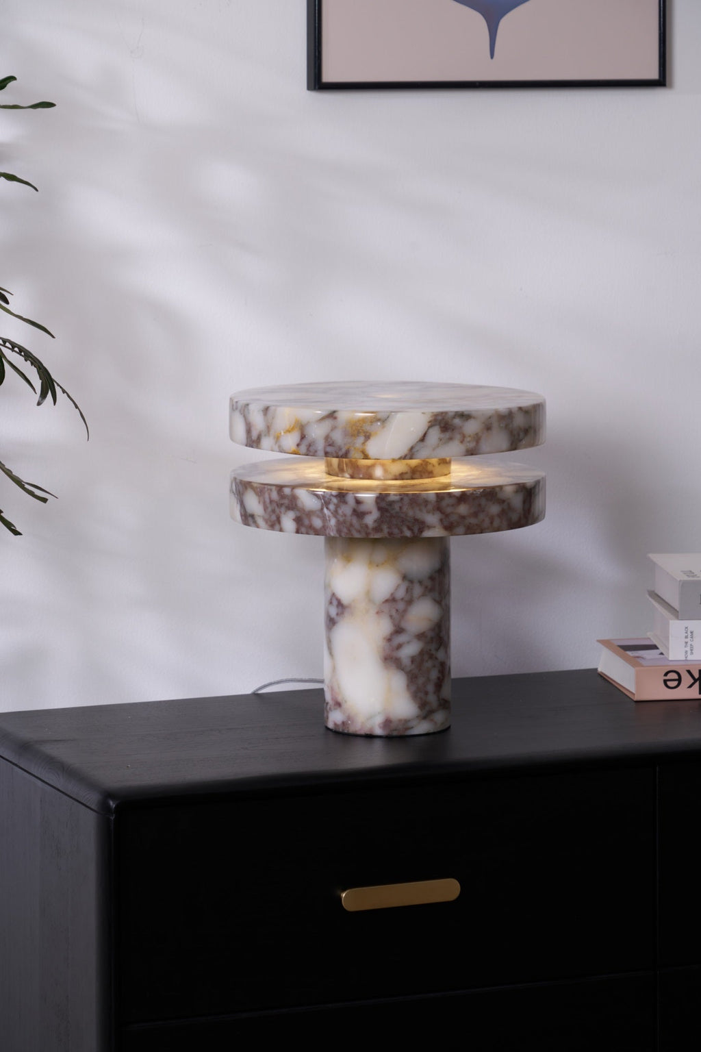Clérachic lamp on a dark cabinet beneath a framed print, its marble column base standing beside a small pink book box.