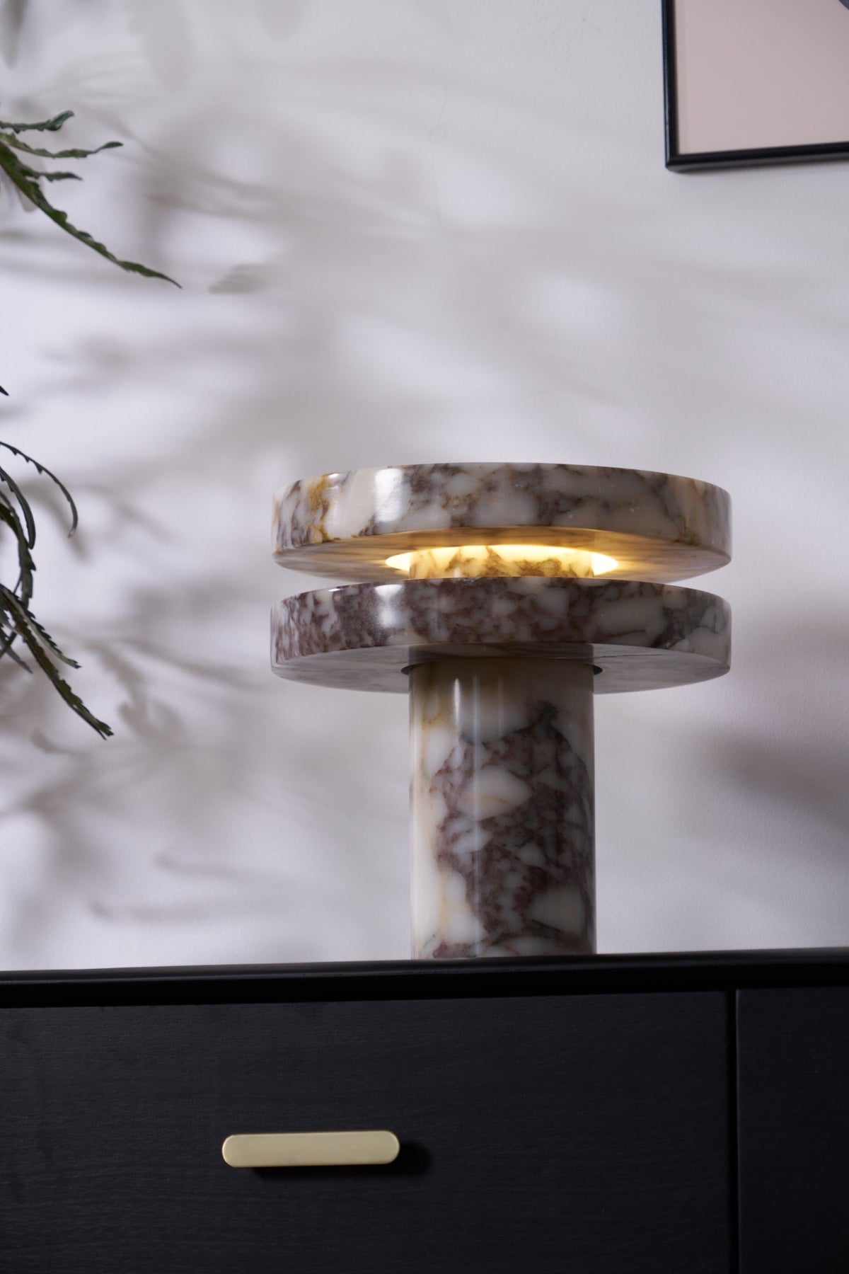Closer crop of the Clérachic lamp on the dark cabinet, a leaf shadow falling across the wall and the marble base.