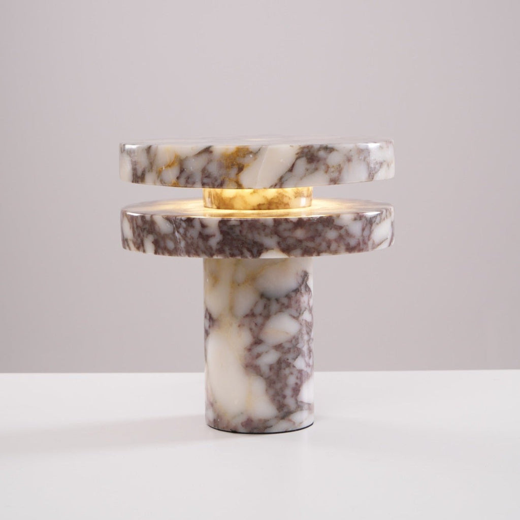 Clérachic lamp lit against a plain pink-beige backdrop, warm light glowing in the gap between its two marble discs.
