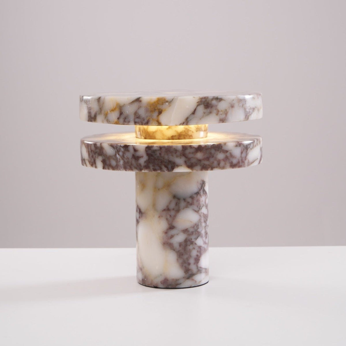 Clérachic lamp lit against a plain pink-beige backdrop, warm light glowing in the gap between its two marble discs.