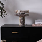 Clérachic lamp lit on a dark dresser, a eucalyptus branch hanging in from the left and stacked books to the right.