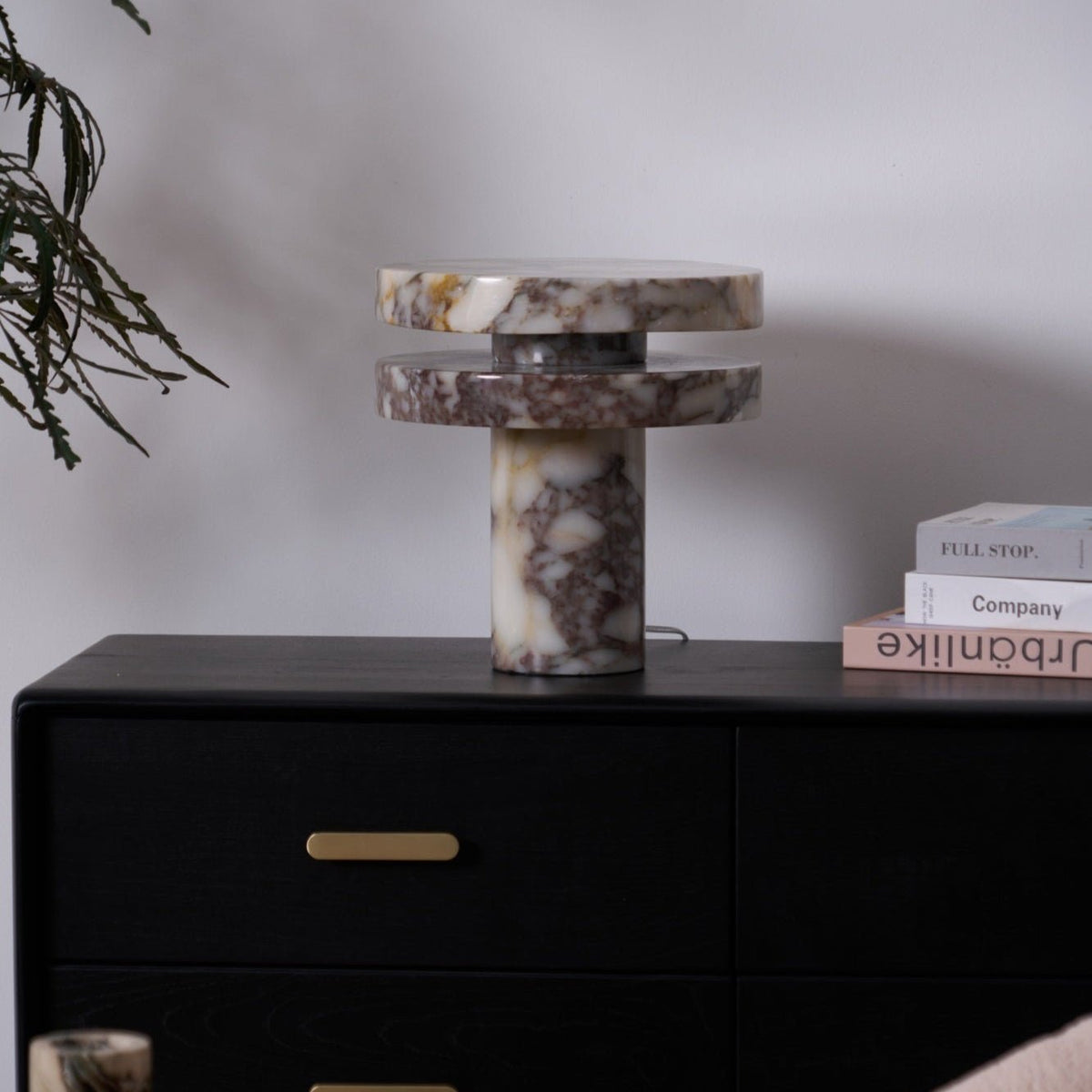 Clérachic lamp lit on a dark dresser, a eucalyptus branch hanging in from the left and stacked books to the right.