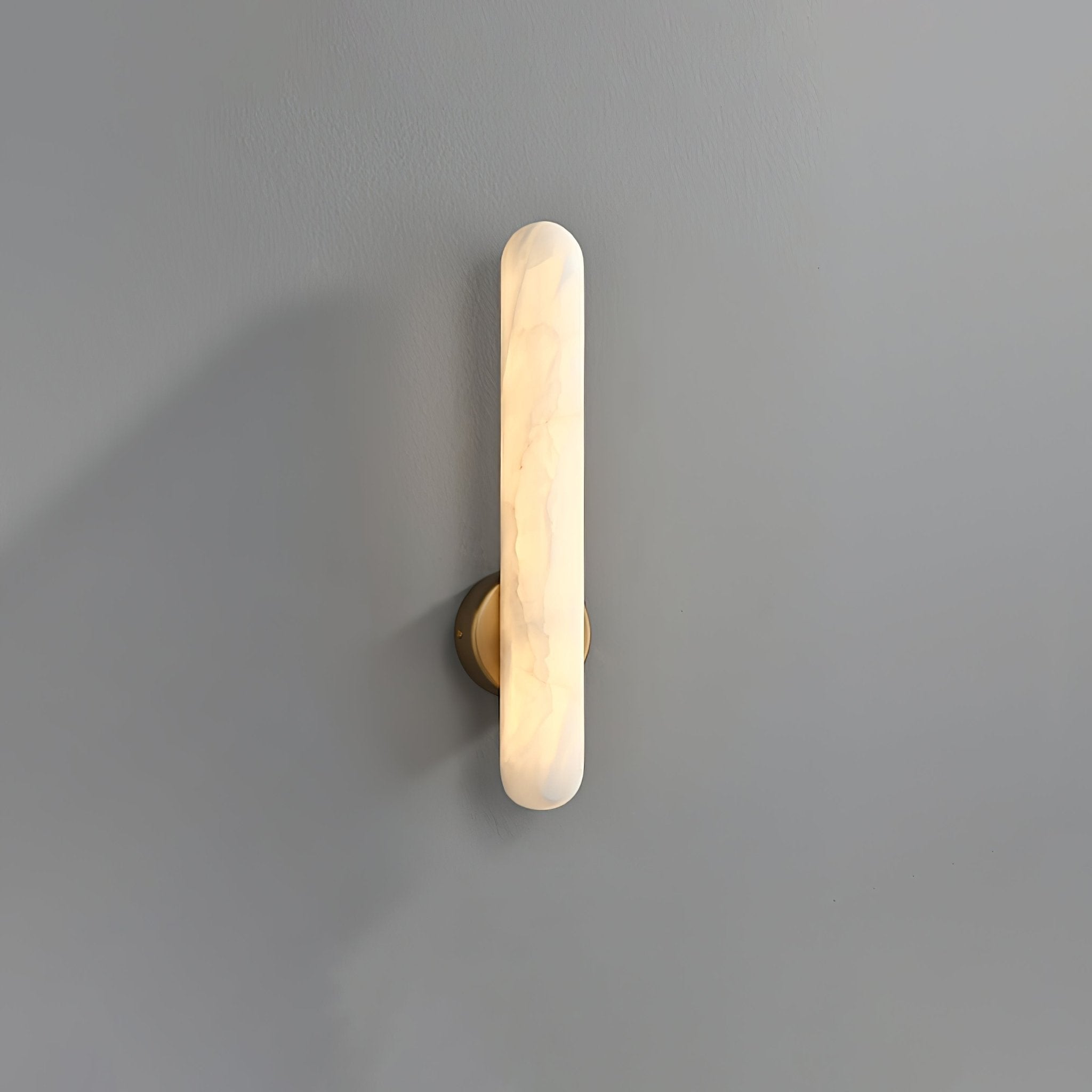 Alabaster bar wall light lit on a plain grey wall, its brass mounting disc centred on the stone.