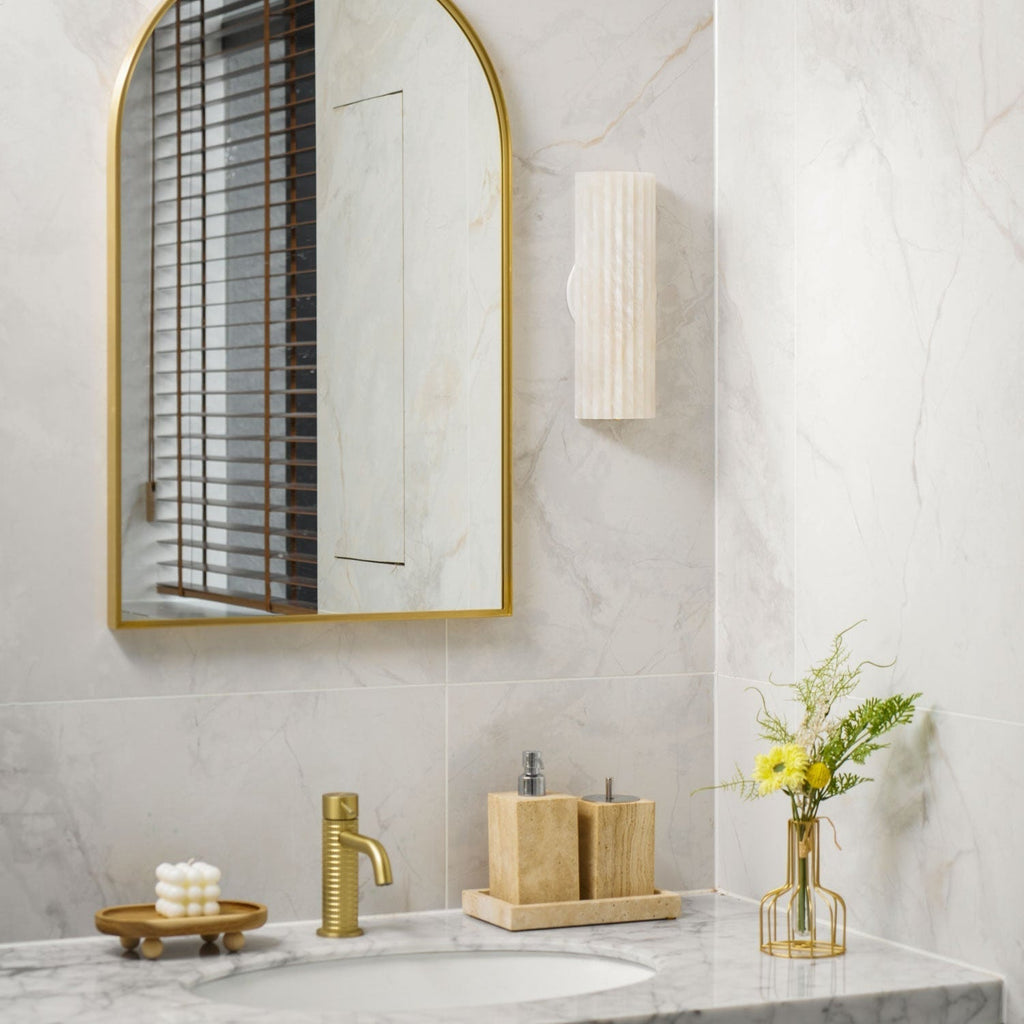 Orcarion alabaster wall light unlit beside an arched brass mirror in a marble-tiled bathroom.