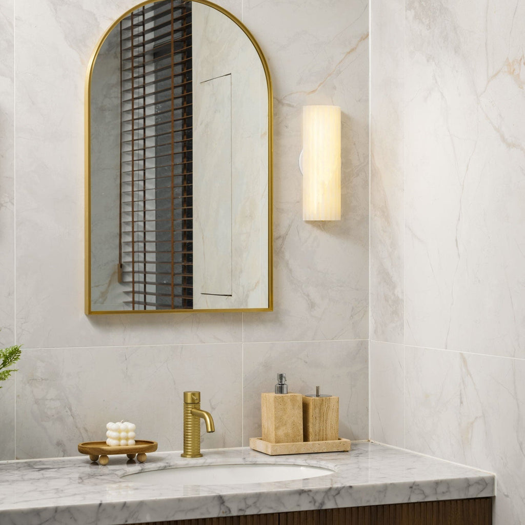 Orcarion alabaster wall light lit beside an arched brass mirror, casting warm light across marble tile.