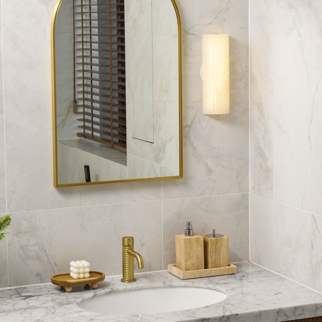 Warm glow of the lit Orcarion wall light above a marble vanity with a brass tap and travertine holders.