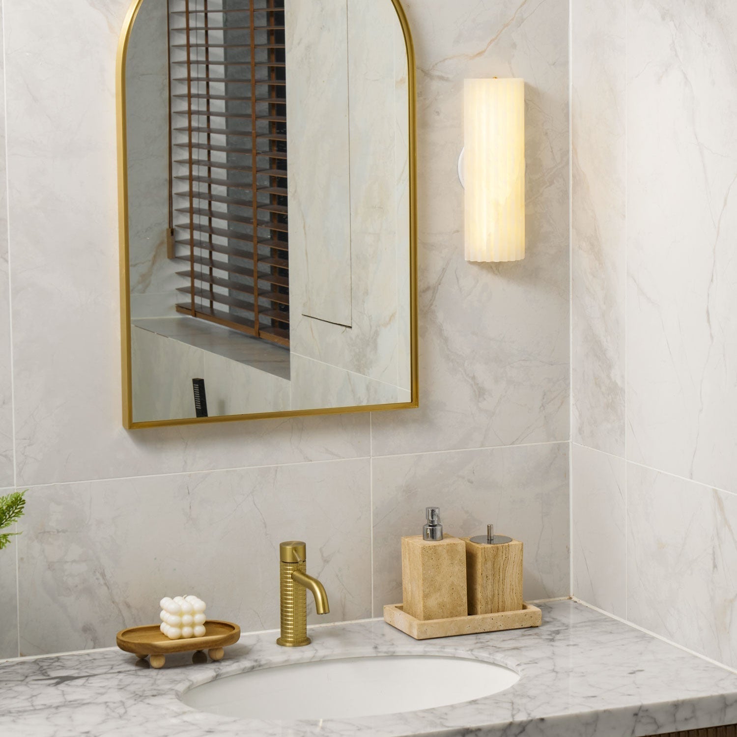 Warm glow of the lit Orcarion wall light above a marble vanity with a brass tap and travertine holders.