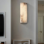 Orcarion alabaster wall light lit in a hallway beside a doorway, with framed art resting below it.