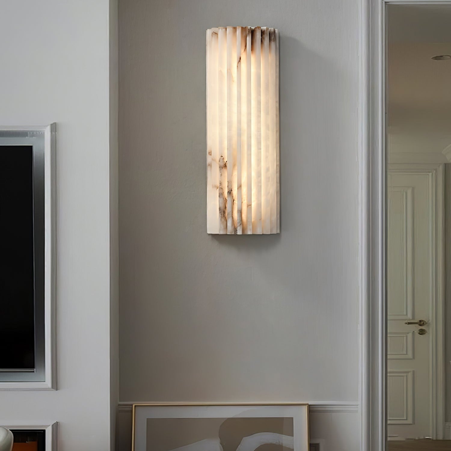 Orcarion alabaster wall light lit in a hallway beside a doorway, with framed art resting below it.
