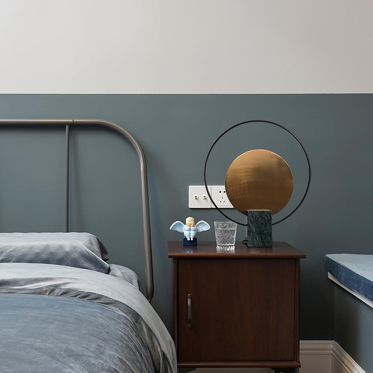 Clérachic table lamp on a wooden nightstand next to an industrial metal-frame bed, a small figurine and glass beside it.