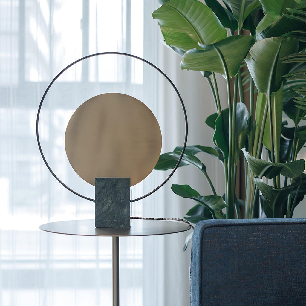 Close-up of a Clérachic table lamp's gold disc and black ring, tall green plant leaves filling the background behind it.
