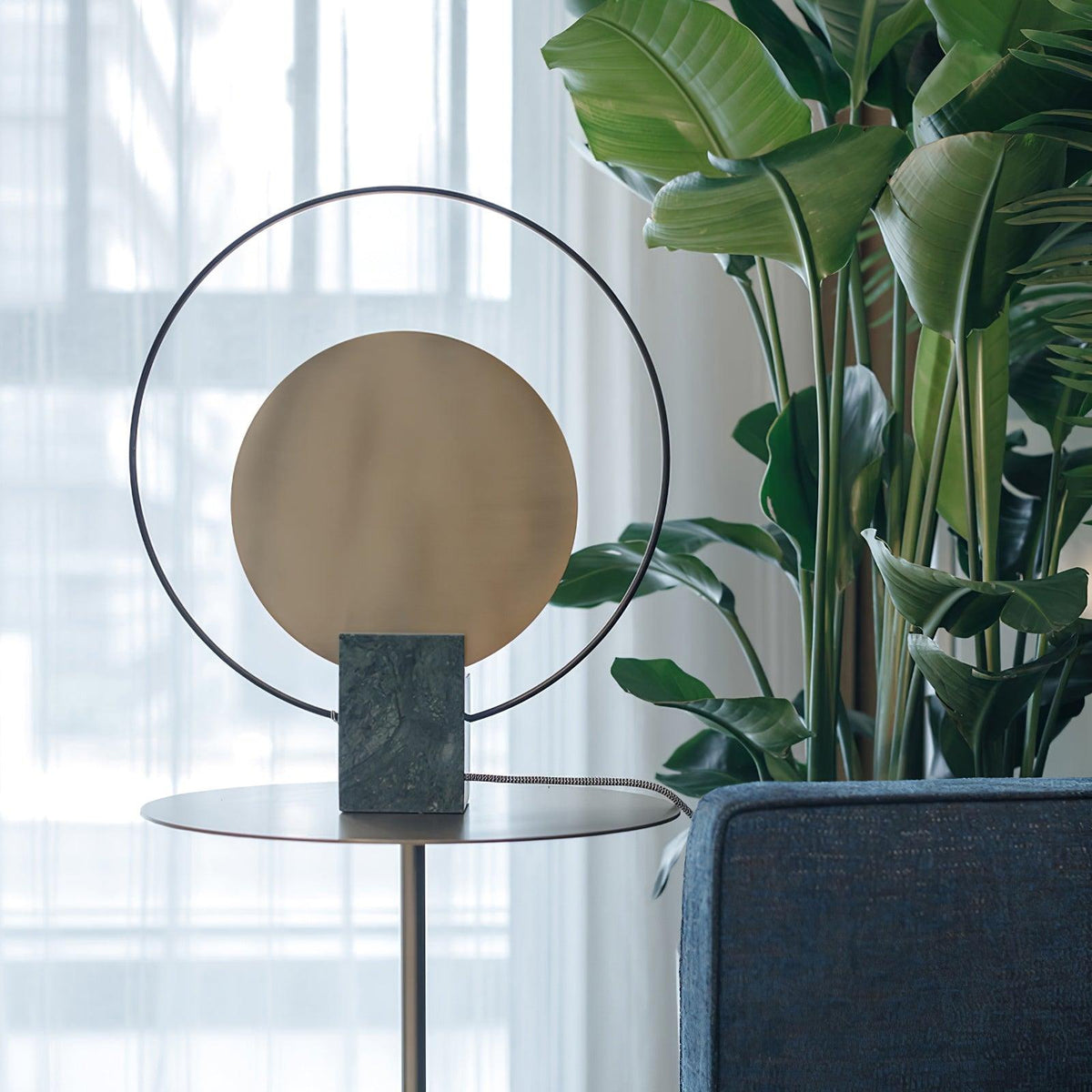Close-up of a Clérachic table lamp's gold disc and black ring, tall green plant leaves filling the background behind it.