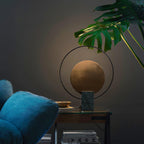 Clérachic table lamp on a side table beside a teal velvet armchair, a large monstera leaf reaching over it from above.