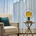 Clérachic table lamp on a glass-and-brass side table next to a beige armchair, sheer curtains at the window behind.