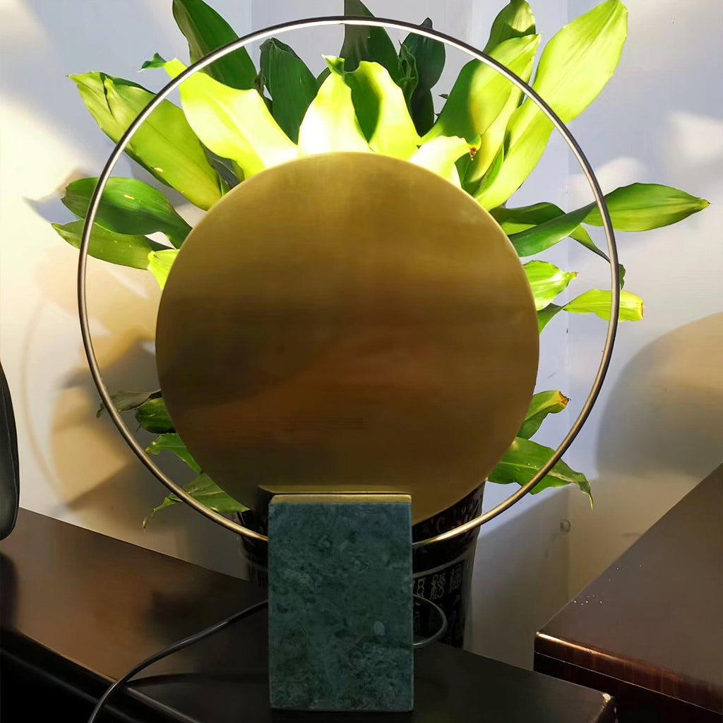 Extreme close-up of a Clérachic table lamp's brushed gold disc, lily leaves framing it above a dark wood tabletop.