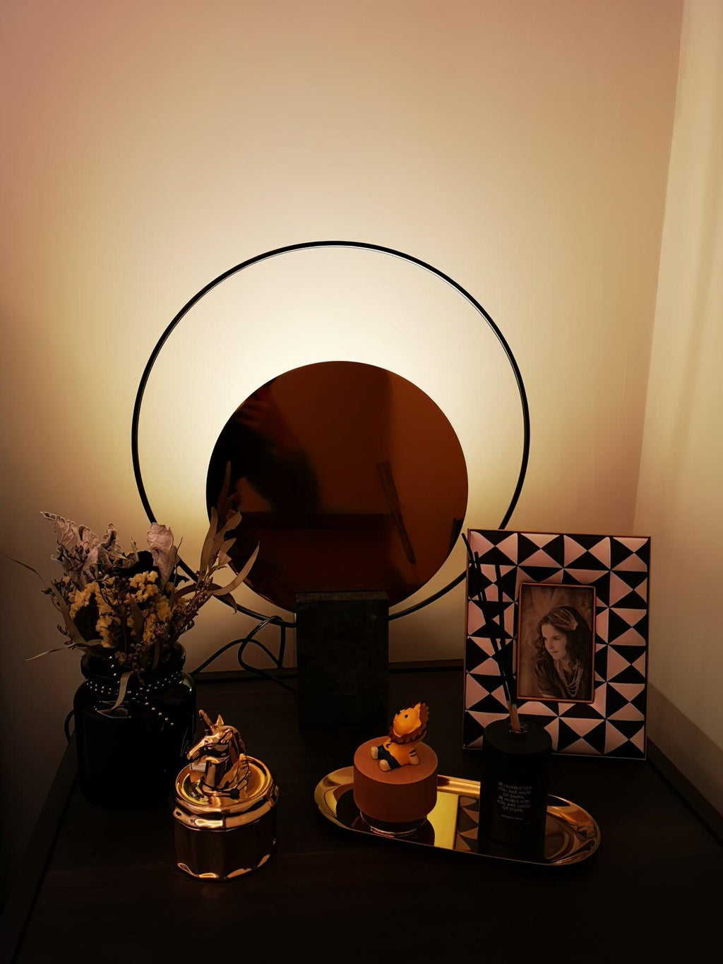 Clérachic table lamp glowing at night on a dark nightstand, dried flowers and a patterned photo frame nearby.