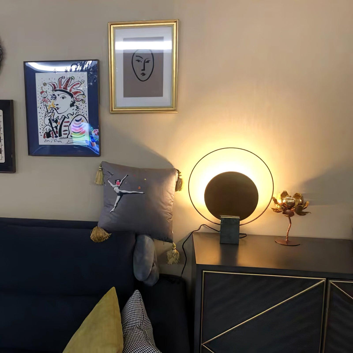 Clérachic table lamp glowing on a glossy black nightstand beside a navy sofa, its gold disc lit warmly in the dim room.