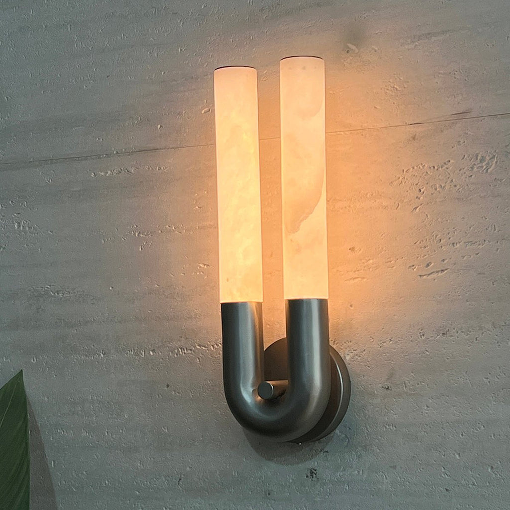 Brushed nickel version of the Arch wall sconce lit at dusk, its twin alabaster tubes glowing