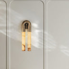 Bronze Arch wall sconce on a white panelled wall in soft daylight, its twin alabaster tubes lit