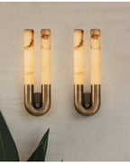 Two bronze Arch wall sconces lit side by side on a pale concrete wall above tropical leaves