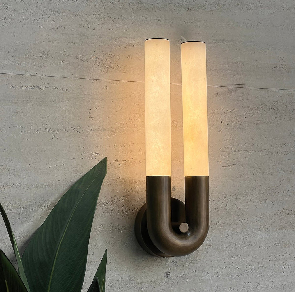 Arch wall sconce in bronze glowing warm on a textured concrete wall beside a green leafy plant