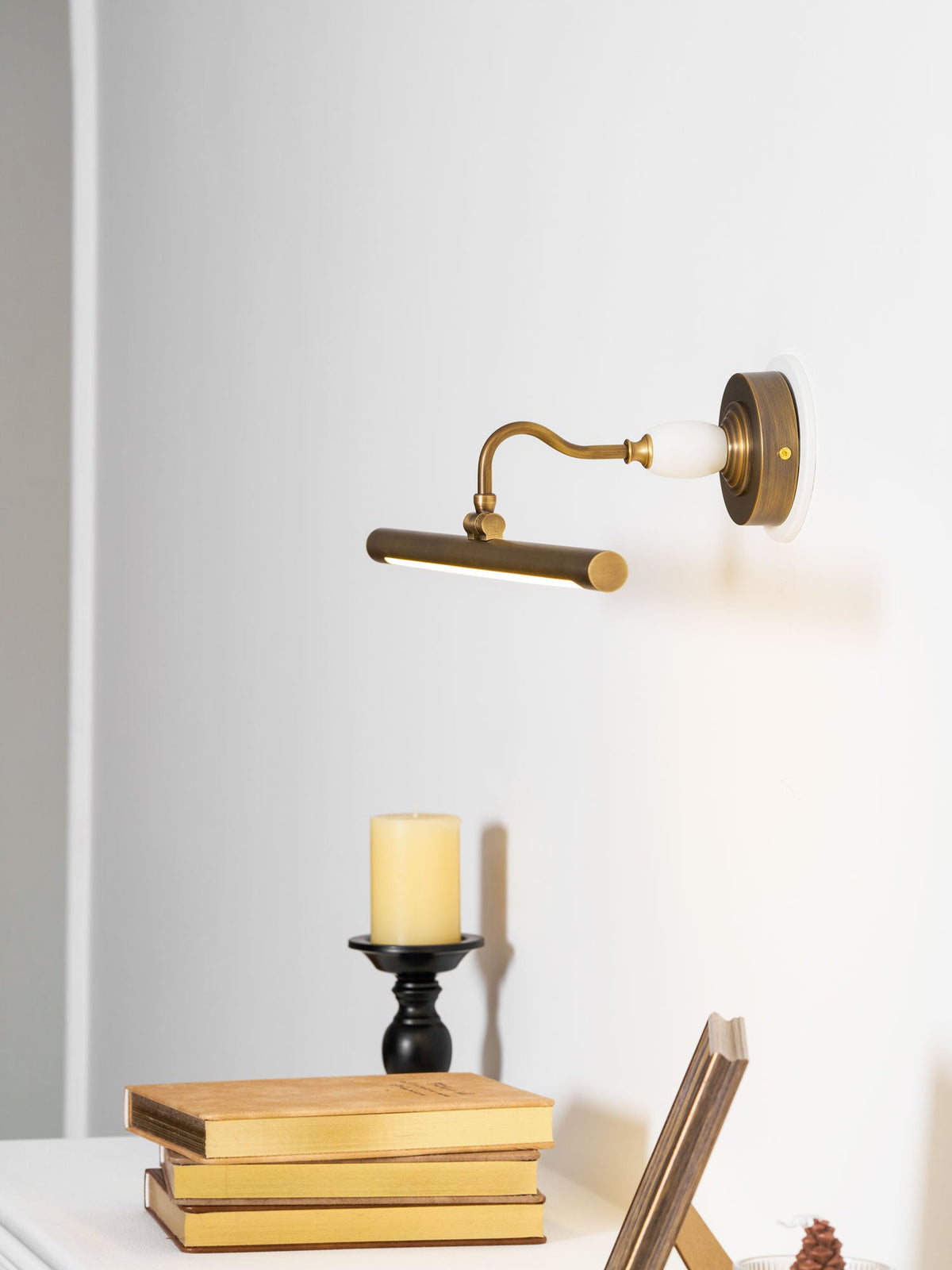 This Clérachic wall sconce in bronze glows above a table with a candle and stacked books, its curved arm visible from the side.