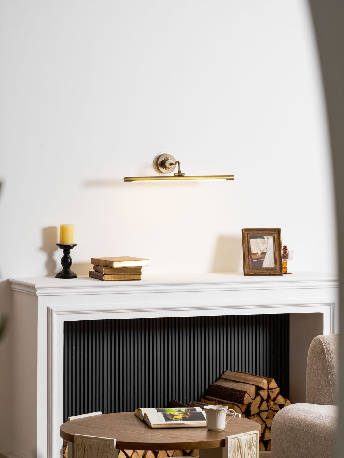 This Clérachic wall sconce in bronze lights a white mantel with a candlestick, books and a framed photo, beside a coffee table.