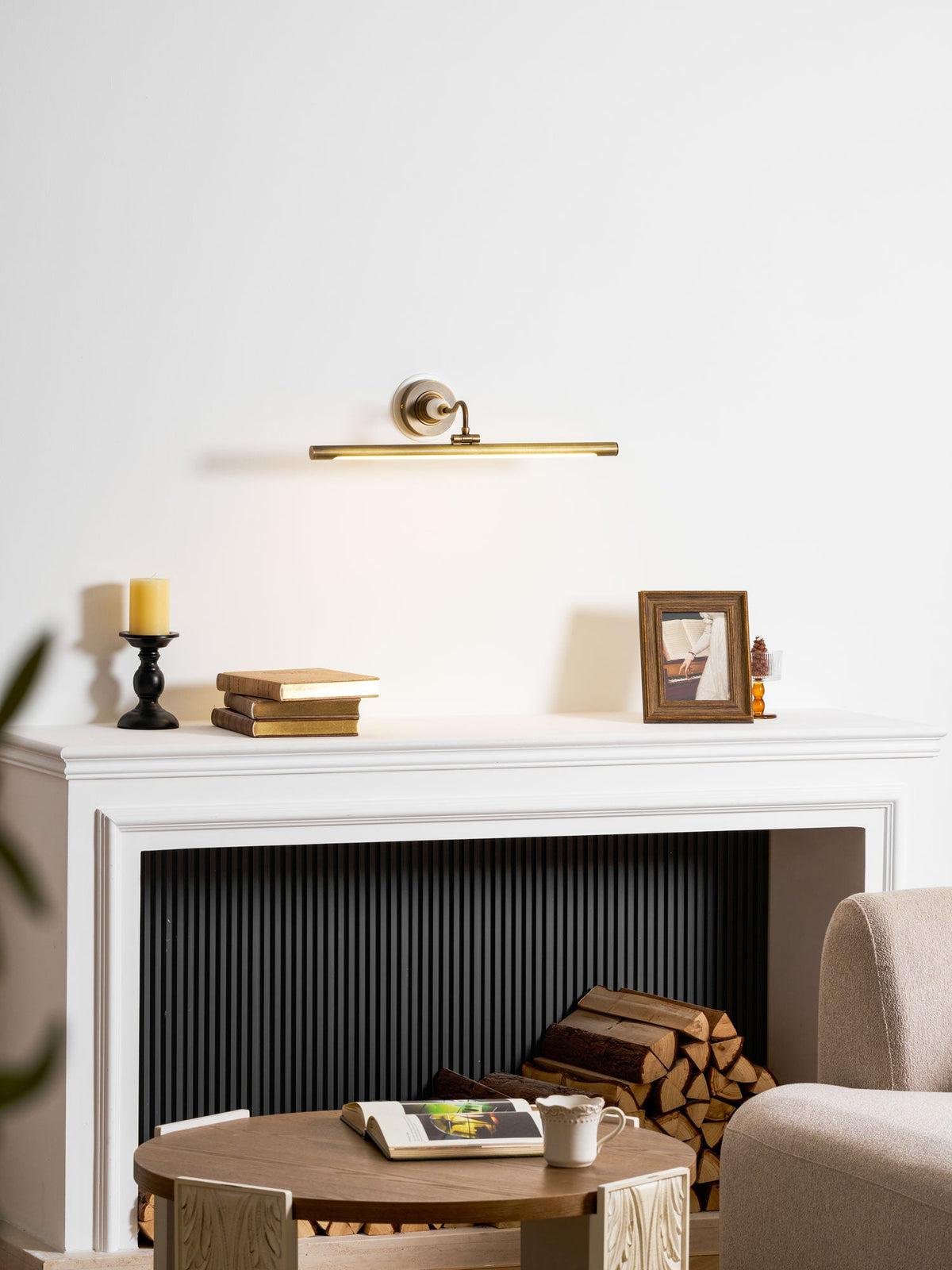 This Clérachic wall sconce in bronze glows above a white mantel with a candlestick and a framed photo, near a log basket.