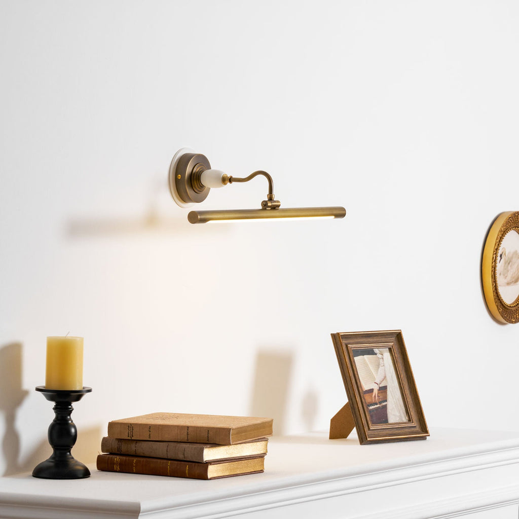 This Clérachic wall sconce in bronze glows above a shelf with a candlestick, books and a framed photo, beside a round wall plate.