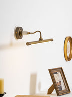 This Clérachic wall sconce in bronze glows above a shelf with a framed photo, near a round decorative plate on the wall.