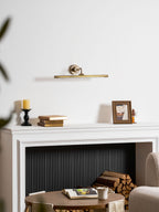 On a white mantel with a candle, stacked books and a framed photo, beside a log basket, sits the glow of this gold Clérachic sconce.