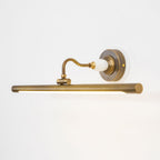 Viewed at an angle, this Clérachic wall sconce in bronze glows along its bar, its curved arm rising from a round wall plate.