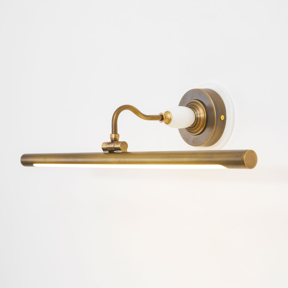 Viewed at an angle, this Clérachic wall sconce in bronze glows along its bar, its curved arm rising from a round wall plate.