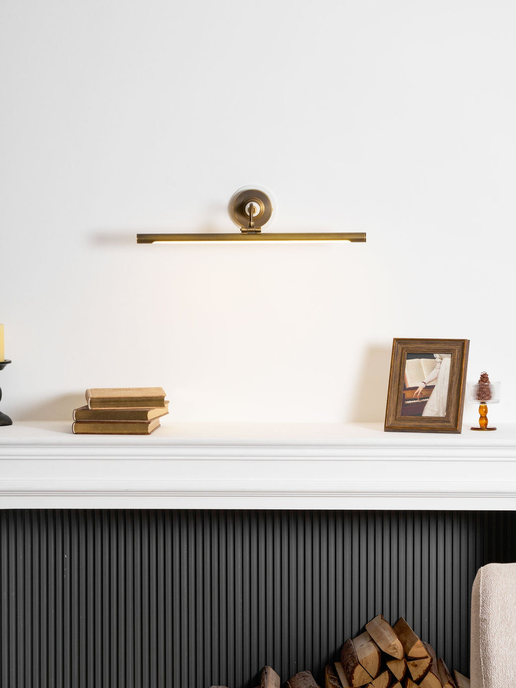Near a dark fireplace surround, a white mantel with stacked books and a framed photo glows under this gold Clérachic wall sconce.