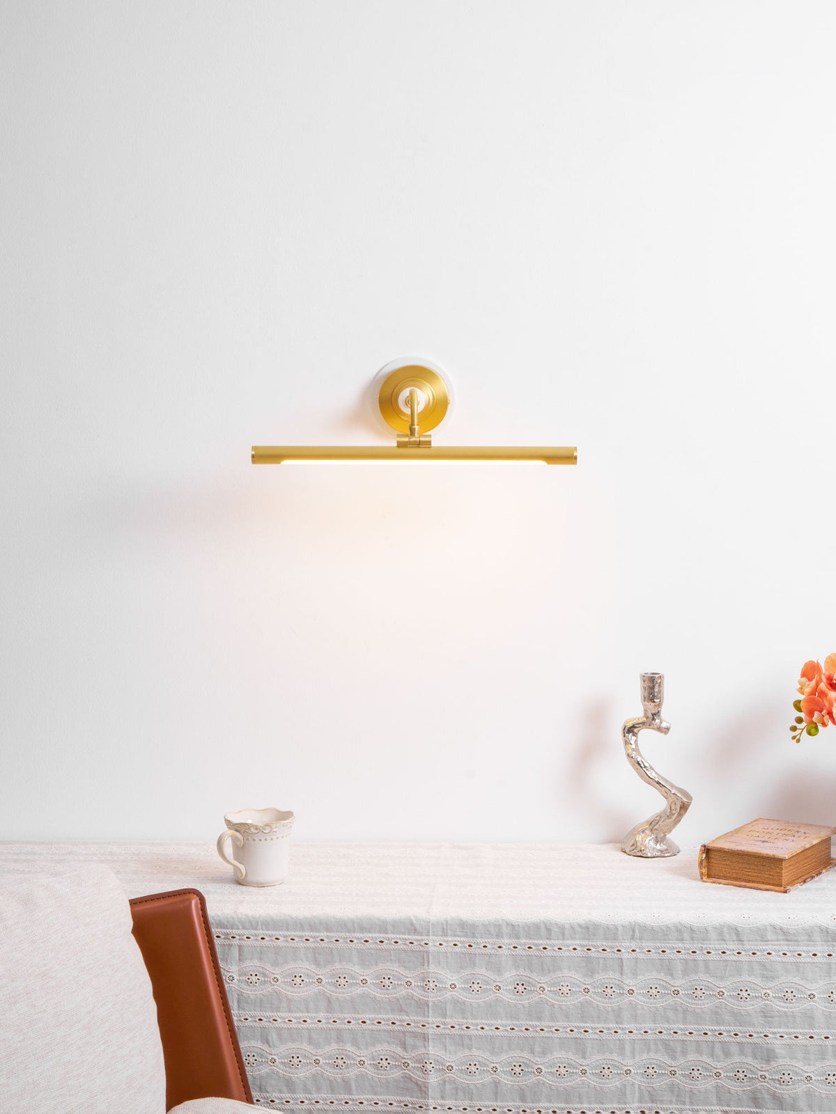 This Clérachic wall sconce in gold lights a plain wall above a table with a jug, candlestick and orange flowers.