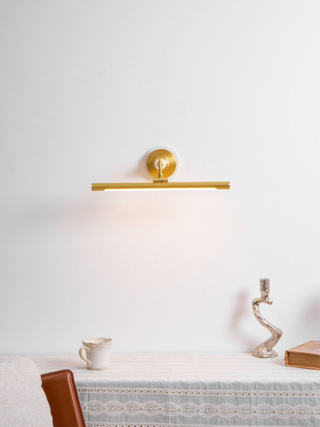 This Clérachic wall sconce in gold glows above a table styled with a jug and candlestick against a plain wall.