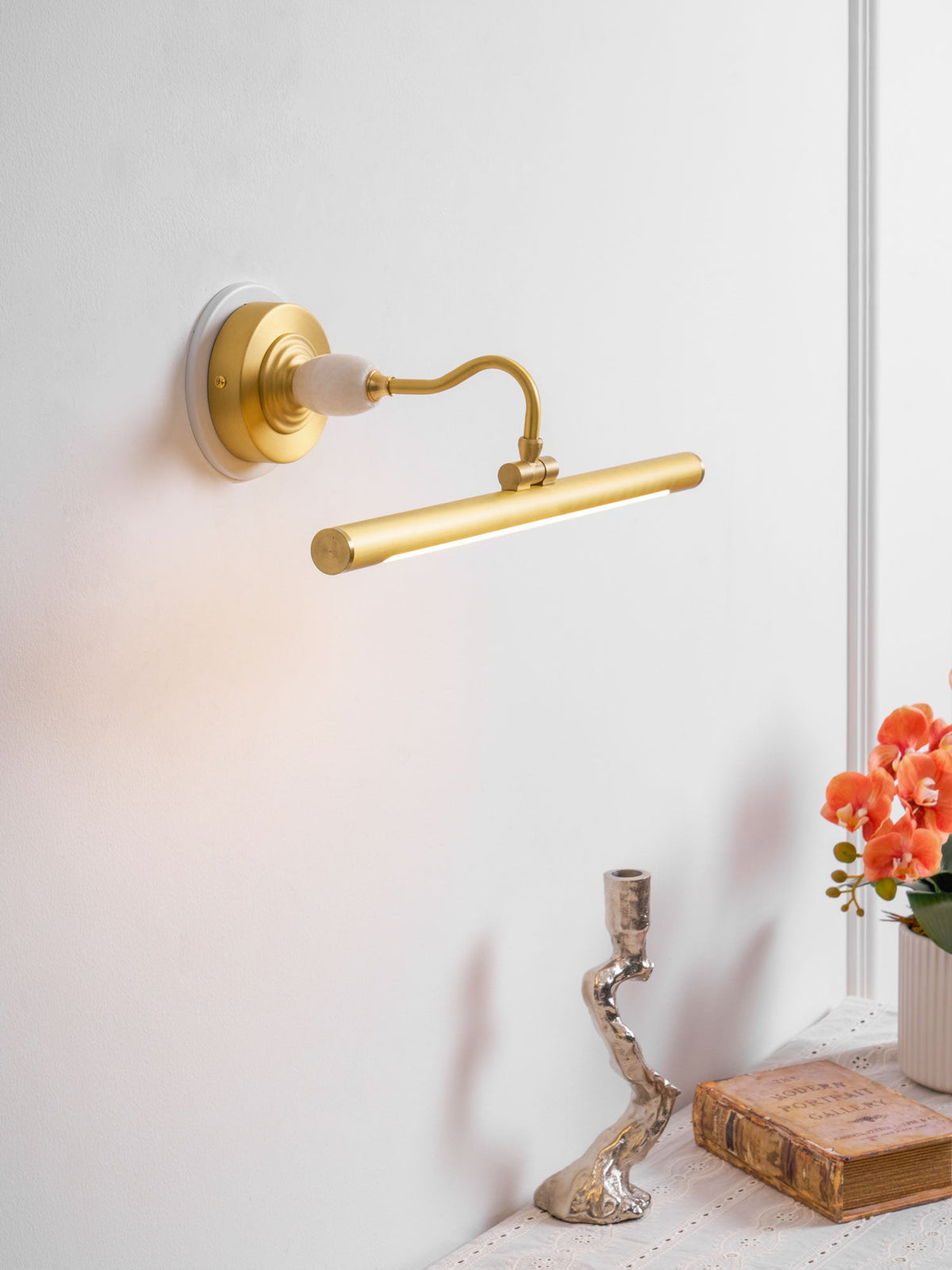 Mounted beside a window, this Clérachic wall sconce in gold glows warmly above a candlestick and a small vase of orange flowers.