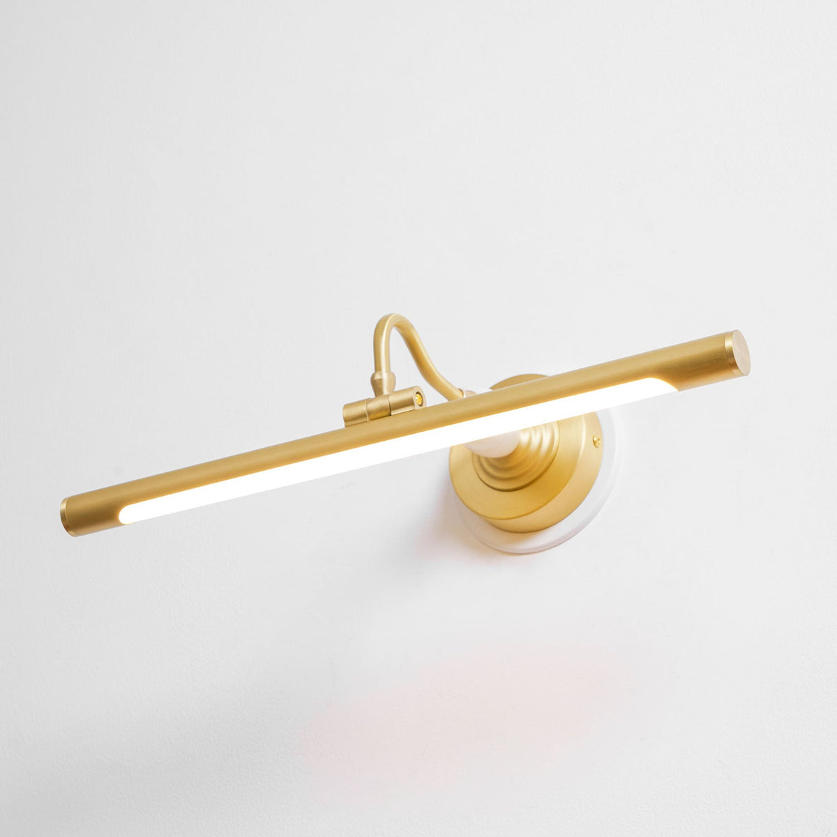 Viewed at an angle, this Clérachic wall sconce in gold glows along its bar, its curved arm connecting to a round wall plate.
