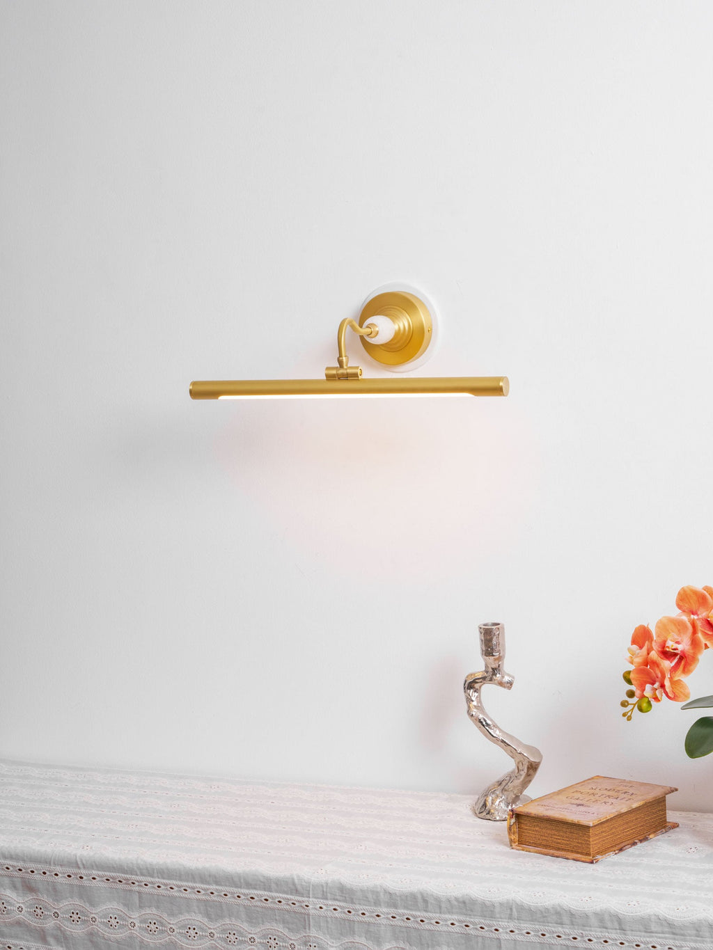 This Clérachic wall sconce in gold glows above a styled table with an ornate candlestick, a book and orange flowers.